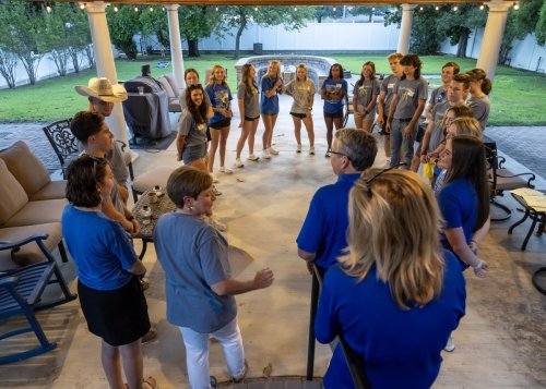A group of 20 diverse individuals stands in front of a white house, wearing blue t-shirts and smiling. 