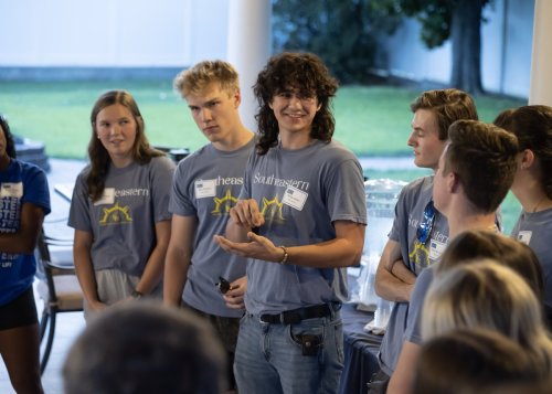 A group of six young adults in casual shirts, engaged in discussion at an outdoor event, with one man speaking confidently in the foreground.