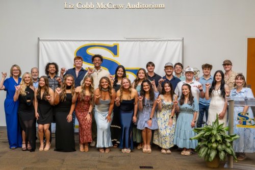 A diverse group of students, approximately twenty-four, celebrates in front of a backdrop with a large blue and yellow S logo in Liz Cobb McCraw Auditorium, many gesturing with shaka signs.