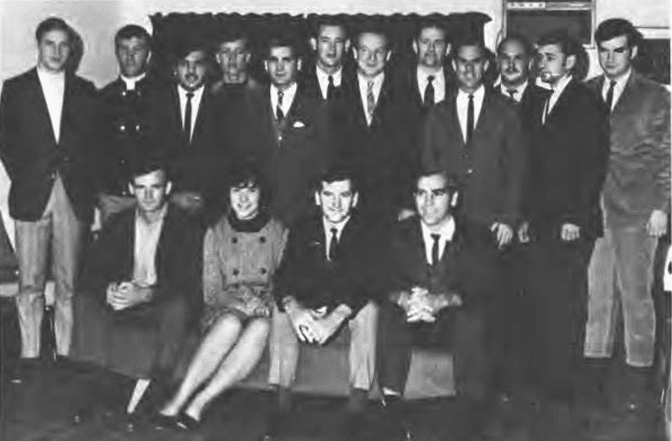A black and white photo of a group of seventeen people, with twelve standing and five seated in front, in formal attire.
