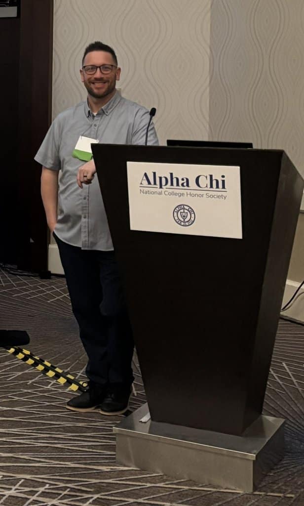 Person standing at a podium with an Alpha Chi National College Honor Society sign.