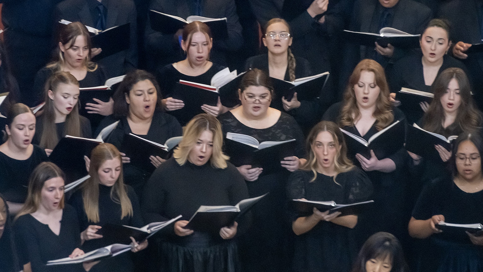 A choir of women singing, holding black music folders, in a formal performance setting.