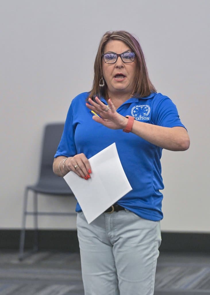 A person in a blue polo shirt with an emblem speaks while holding a paper, gesturing with her other hand.