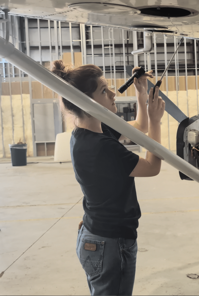 Person inspecting the underside of an aircraft wing with a flashlight and screwdriver in a hangar.
