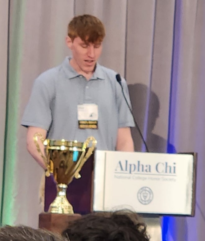 Person standing at a podium with an Alpha Chi National College Honor Society sign.