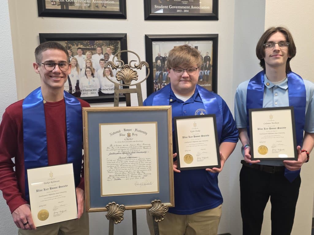 Three young men wearing blue stoles stand in front of a display table with candles, certificates, and group photos on the wall