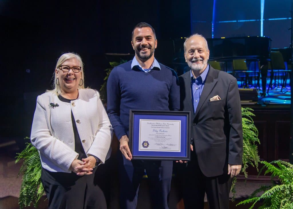 Three people stand together, with the center person holding an award.

