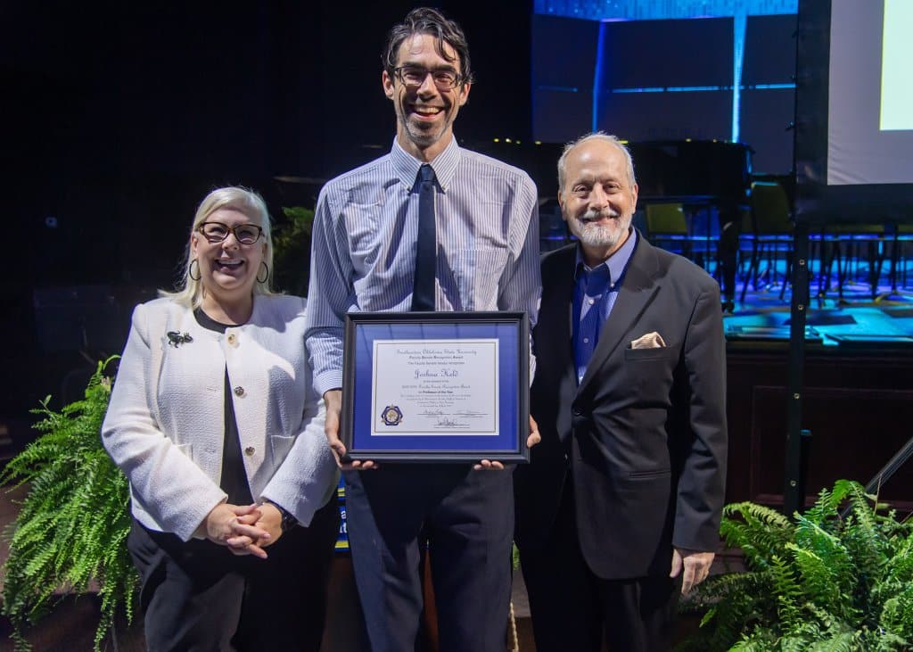 Three people stand together, with the center person holding an award.

