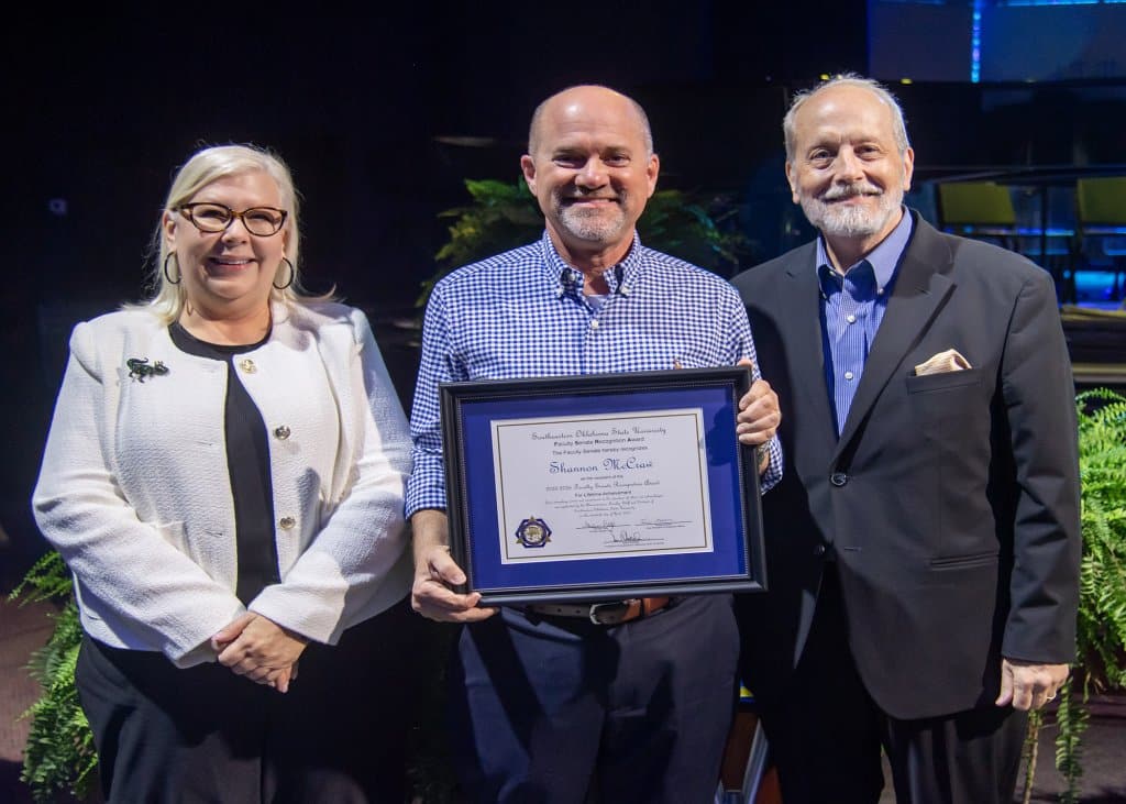 Three people stand together, with the center person holding an award.

