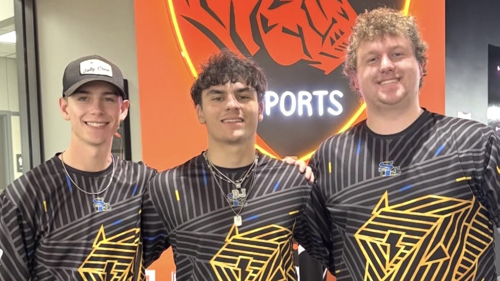 Three people wearing matching black and yellow jerseys stand in front of an orange backdrop with a logo.

