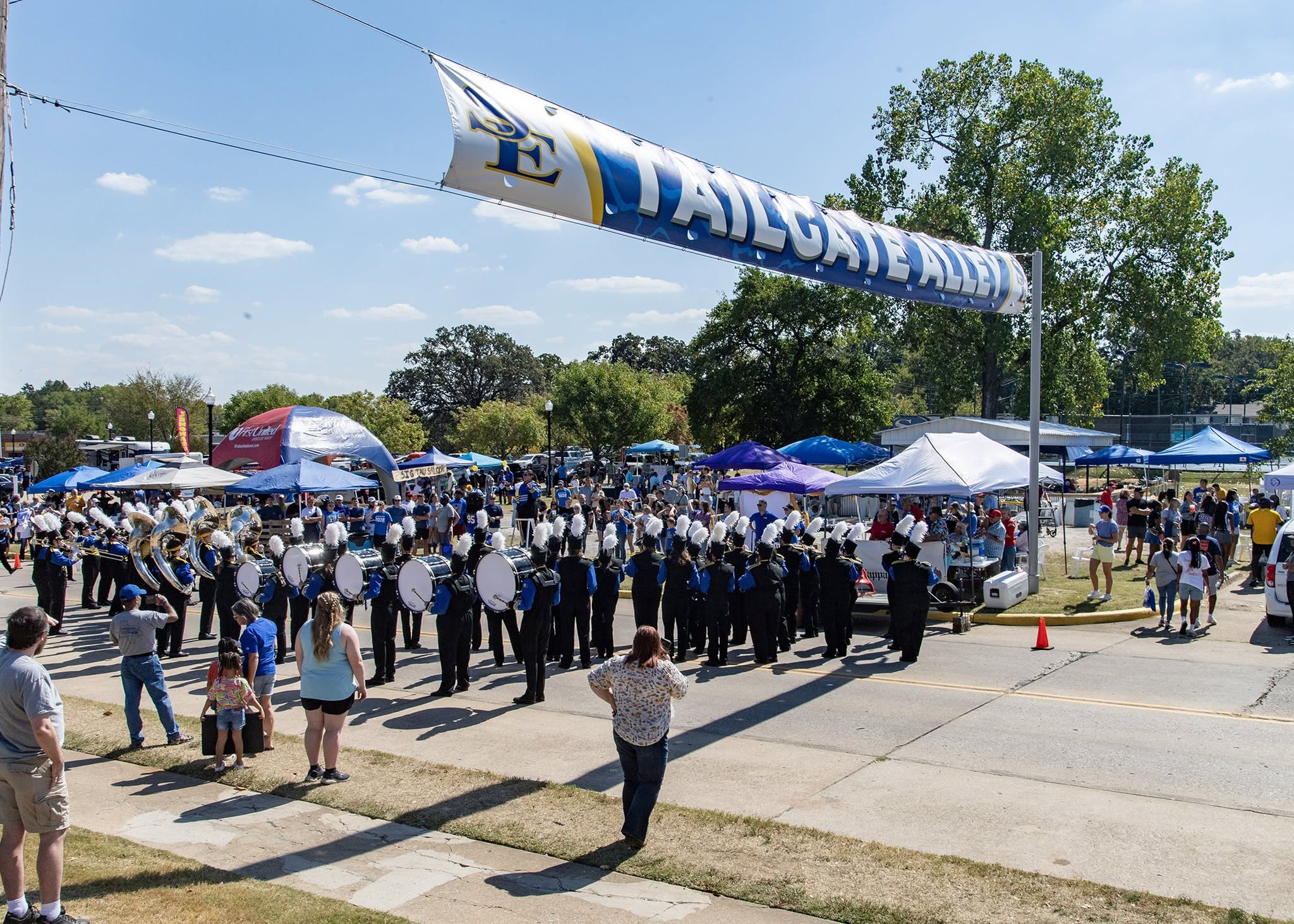 A marching band performs in a street beneath a banner reading “Tailgate Alley,” surrounded by spectators and tents.