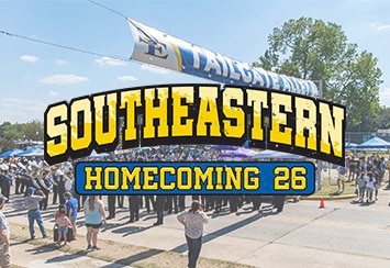 A marching band performs in a street beneath a banner reading “Tailgate Alley,” surrounded by spectators and tents.