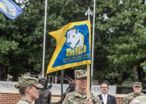 Military personnel in camouflage uniforms with a flag reading GOLD and Oklahoma Army National Guard, Southeastern Oklahoma State University.